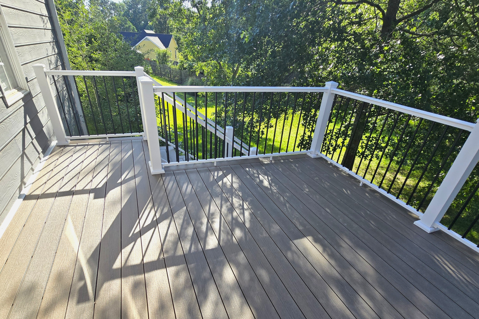 A raised deck with gray composite flooring, white railings, and thin black vertical balusters overlooking a green lawn.