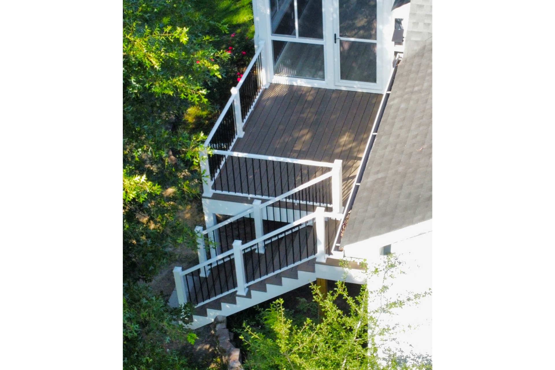 A high-angle view of a white, multi-level wooden deck with brown floorboards and railings, attached to a house near trees.