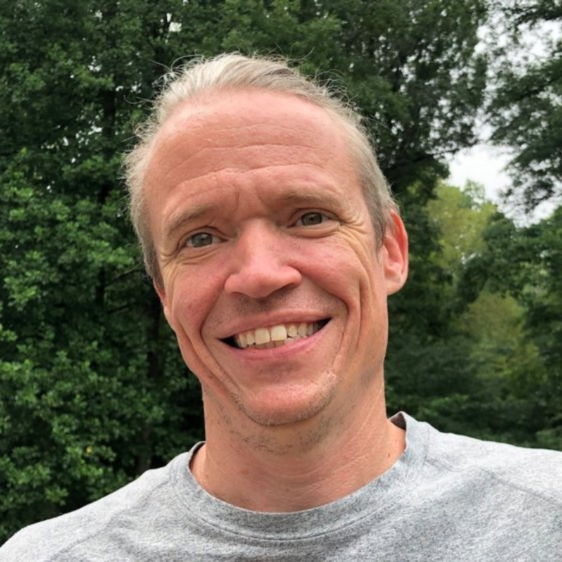 A smiling person with light-colored hair, wearing a gray shirt, against a blurred background of green trees.