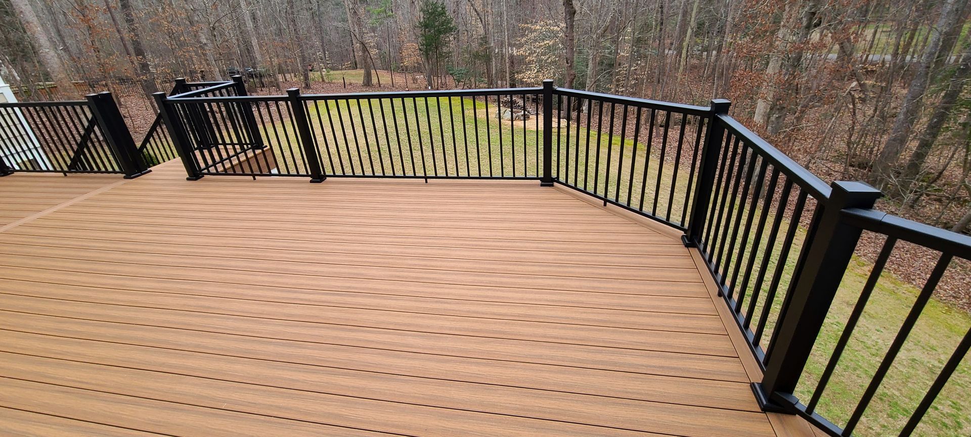 A wooden deck with a black railing, looking out over a grassy yard and a background of bare trees in late autumn.