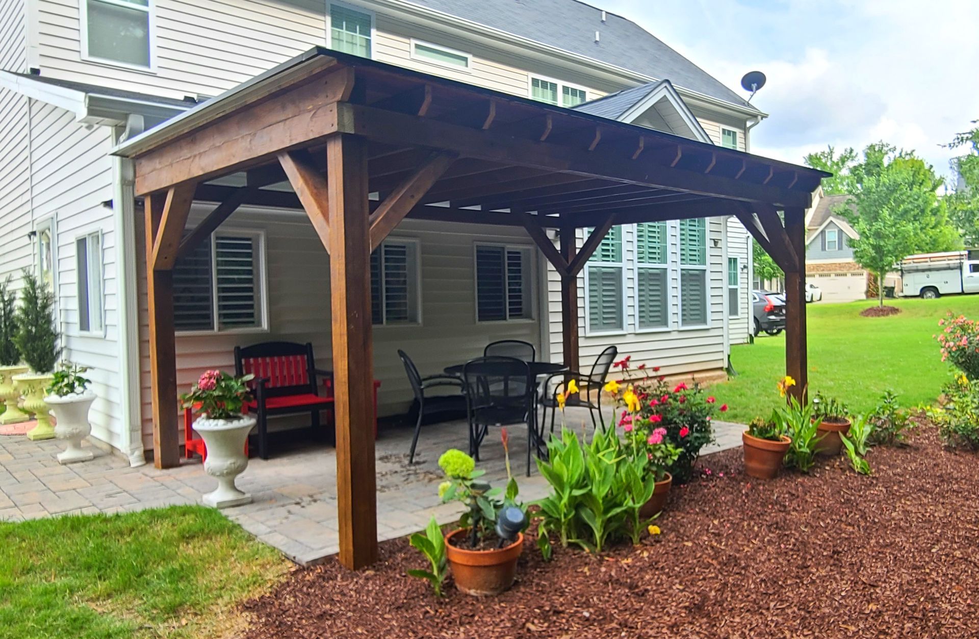 A wooden pergola covers a stone patio with a table, chairs, and bench, situated next to a house with a landscaped garden.