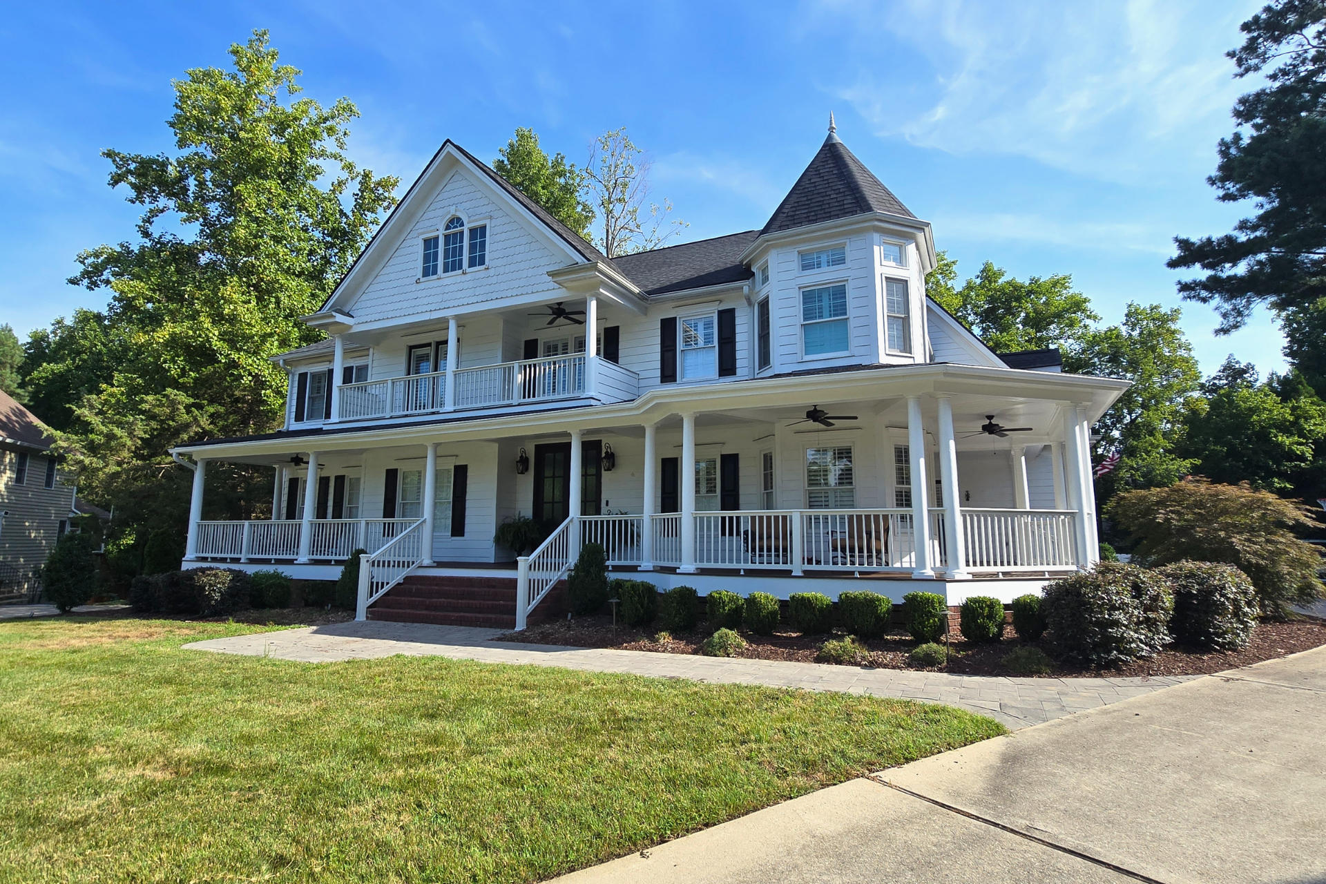 A two-story white Victorian-style house with a wraparound porch, a corner turret, and a lawn under a bright blue sky.