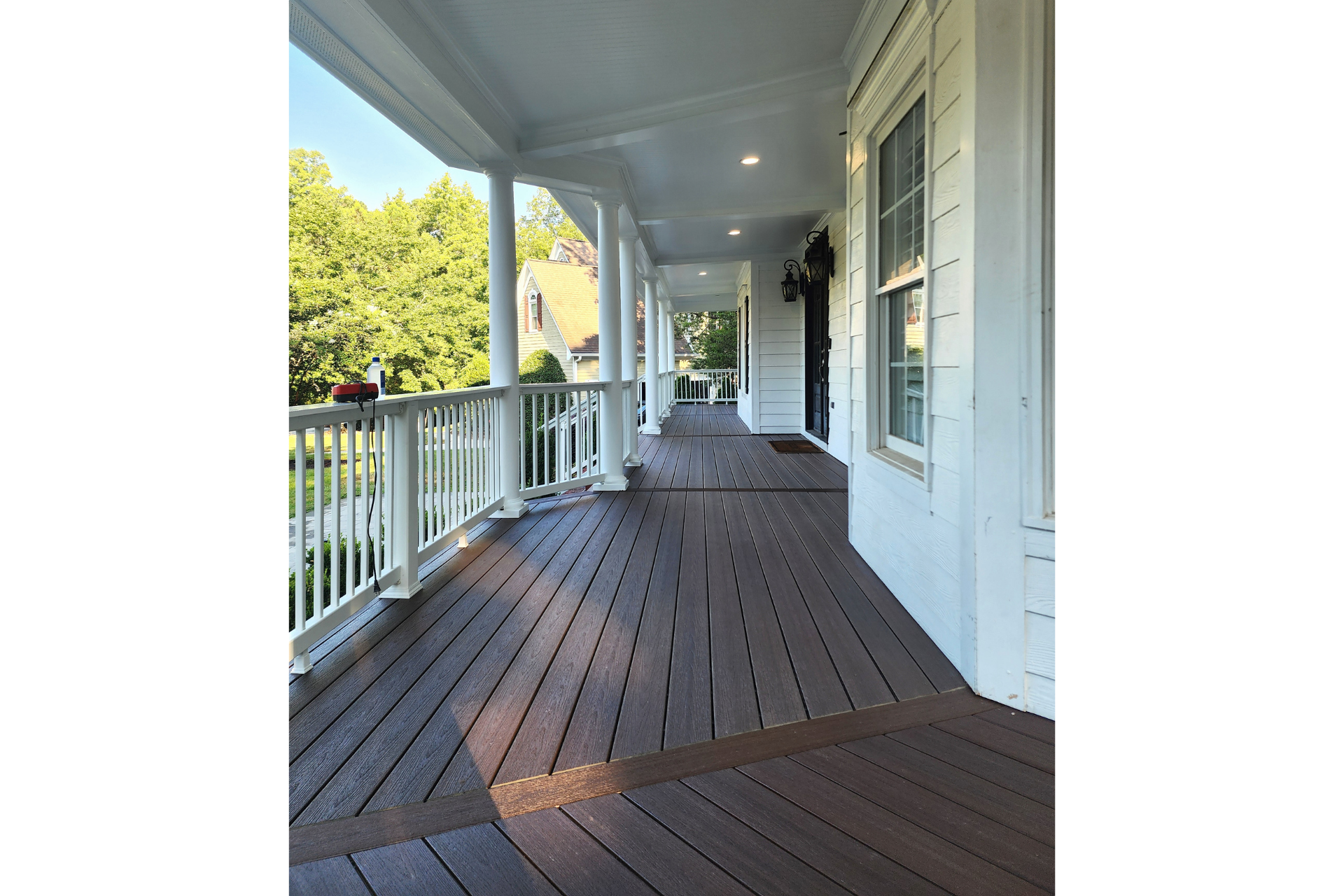 A wide, covered porch with dark composite decking, white columns, and a white railing overlooking a sunny, green yard.