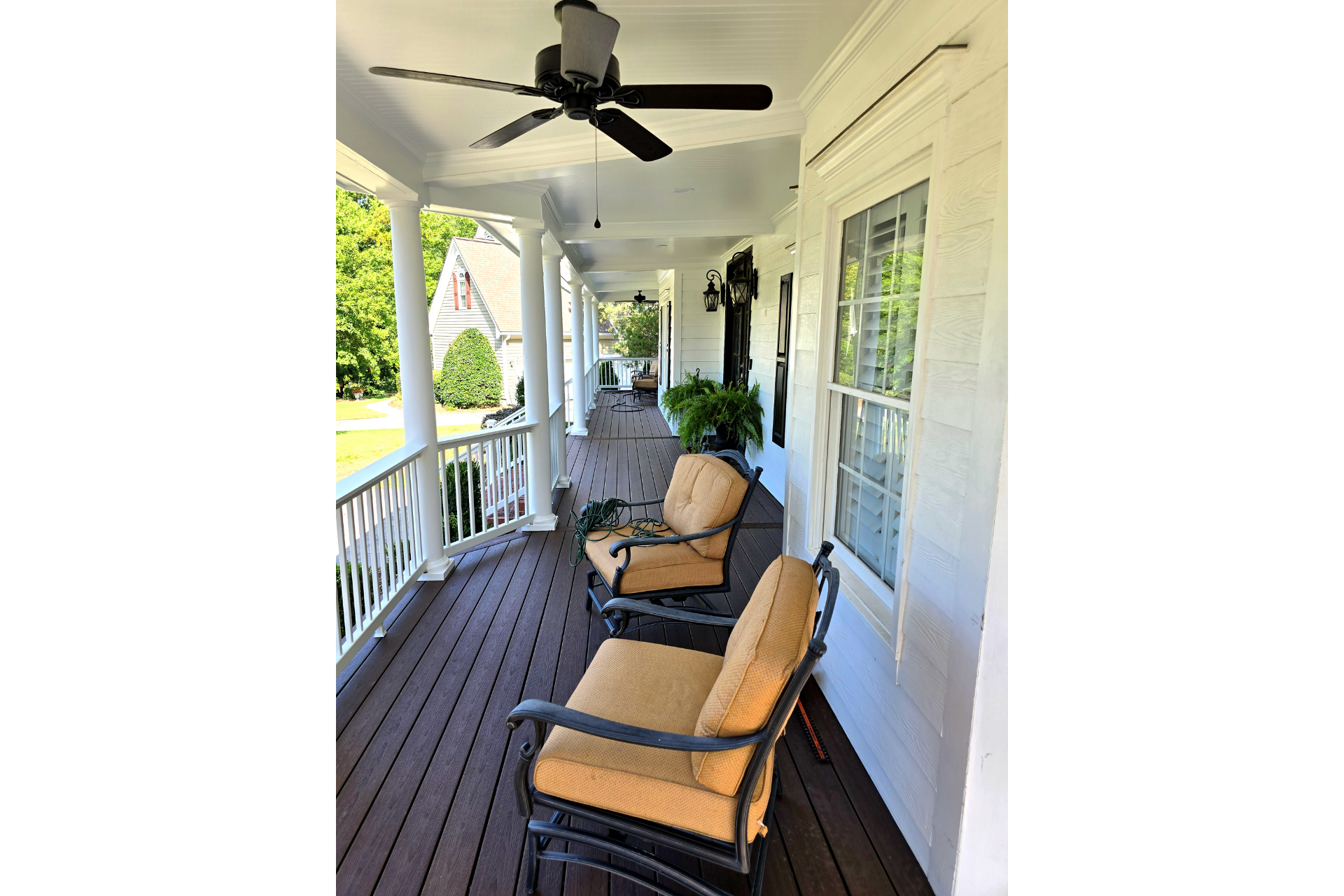 A covered porch with white columns, brown flooring, and two beige-cushioned armchairs, looking toward a sunny lawn.