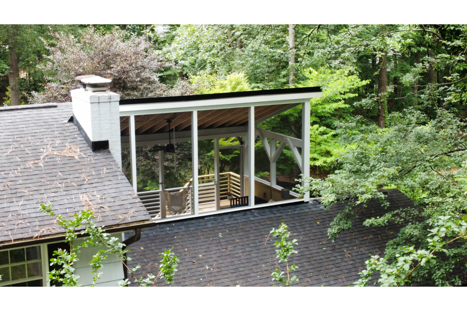 An elevated outdoor covered porch attached to a shingled roof house, surrounded by lush green trees.