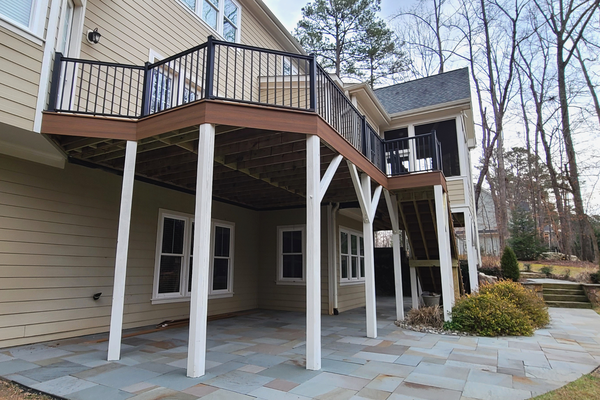 A raised wooden deck with black railings and stairs over a stone patio against a tan-sided house in a wooded setting.