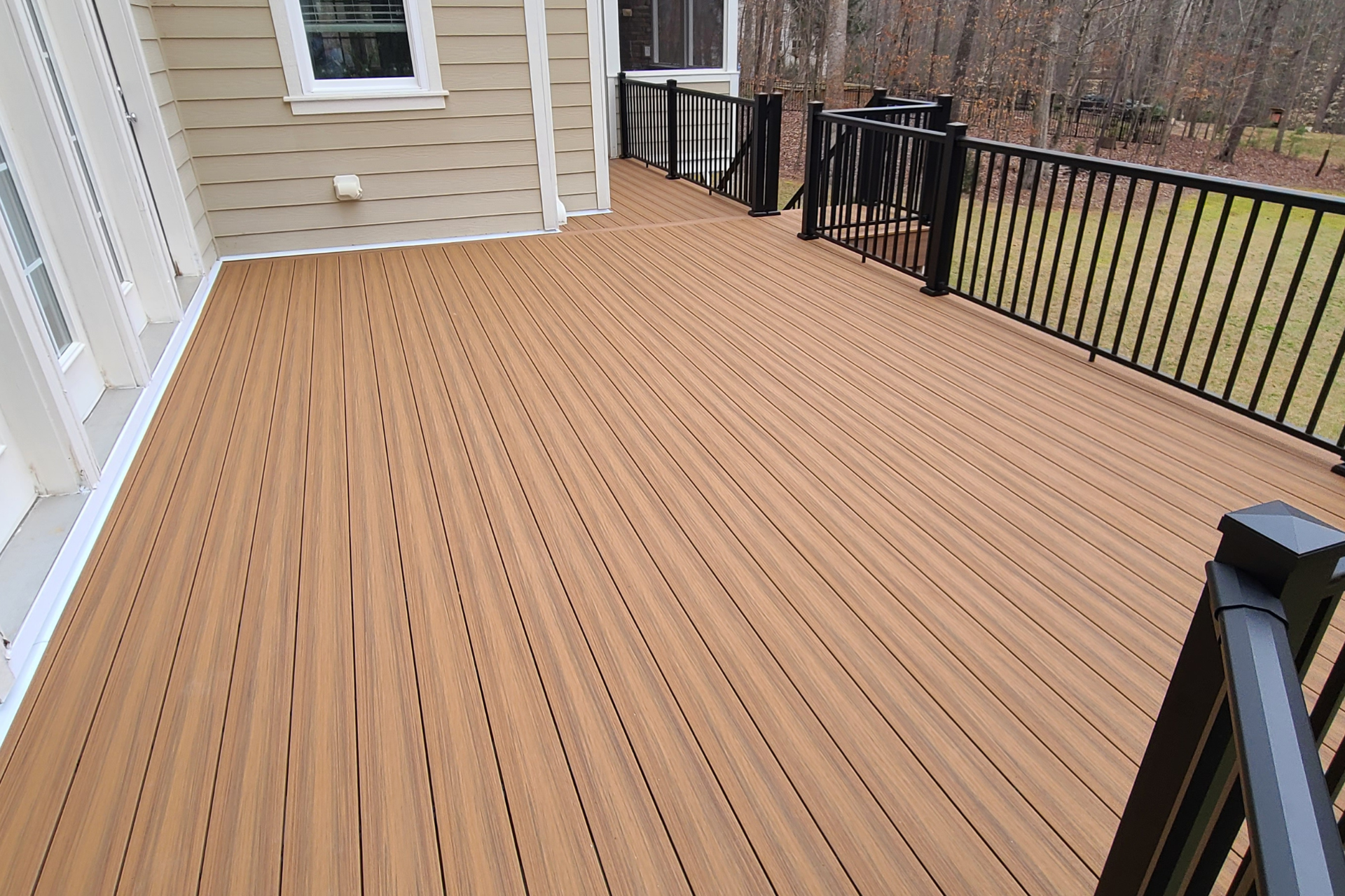 A tan, composite-plank deck with black railings extends from a light-colored house toward a grassy backyard.