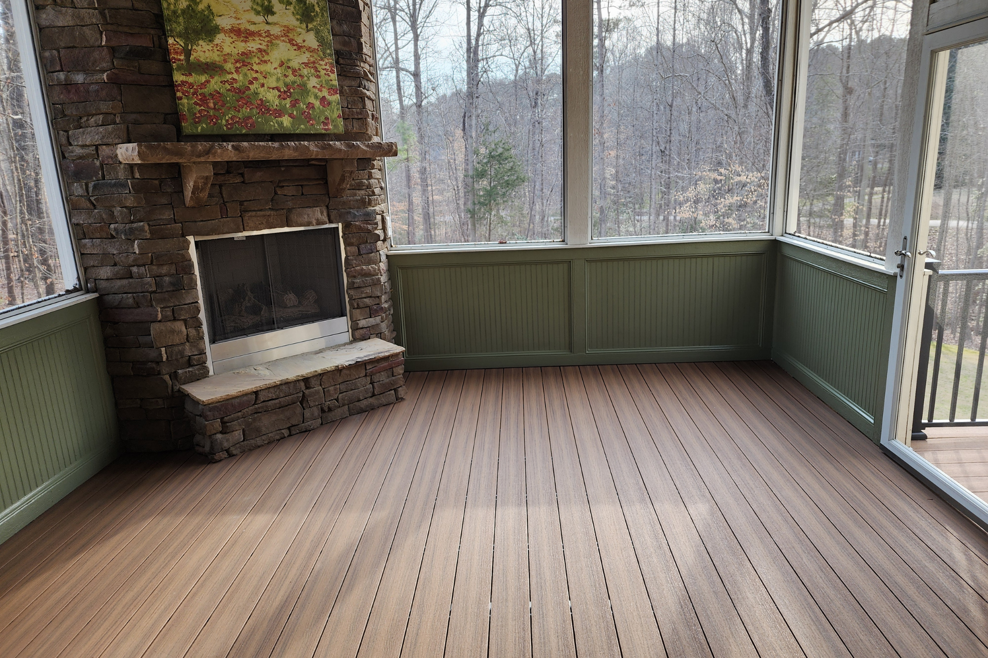 A screened-in porch with a stone fireplace, light brown composite wood flooring, and green wainscoting overlooking trees.