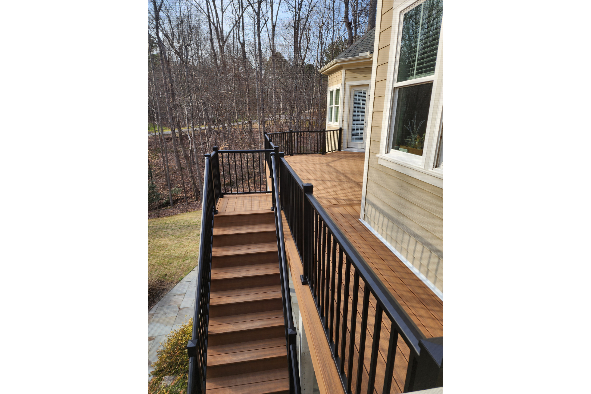 A wooden deck with black railings and stairs leading down to a grassy area next to the side of a house.