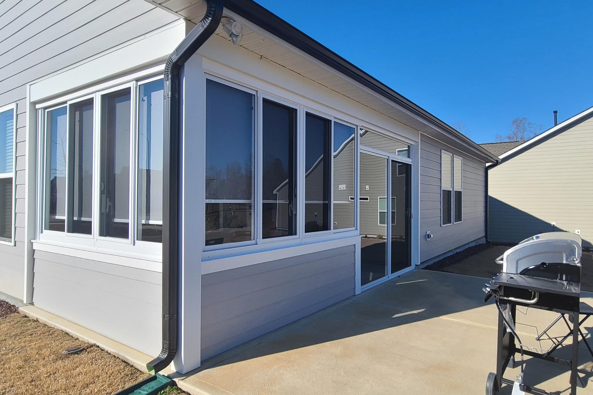 Light gray home exterior featuring a sunroom with large windows, a sliding glass door, and a concrete patio with a grill.