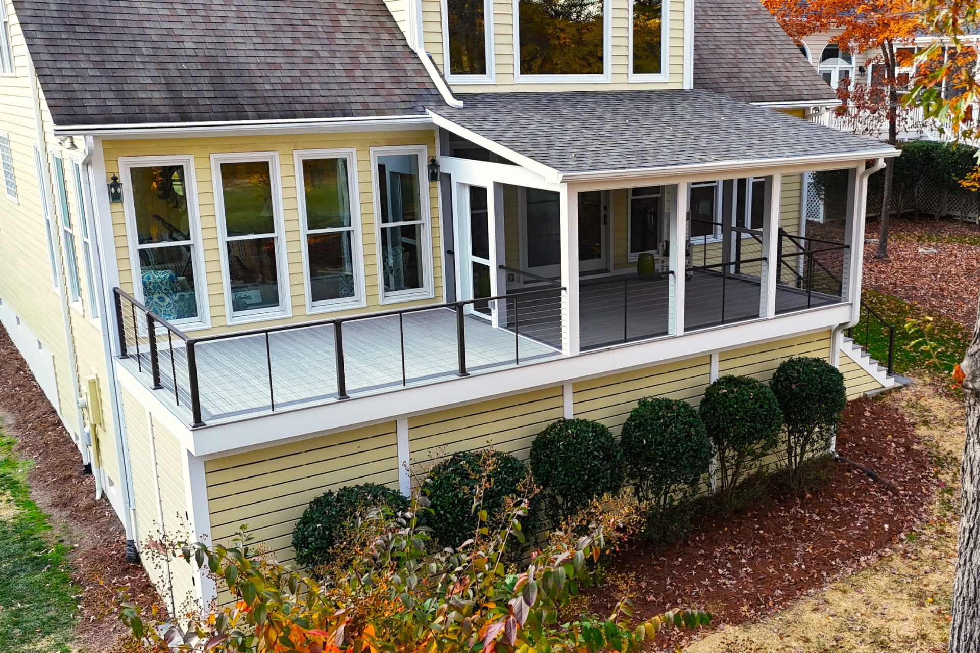 A yellow two-story house with a white-railed deck and a covered, screened-in porch, overlooking a landscaped garden.