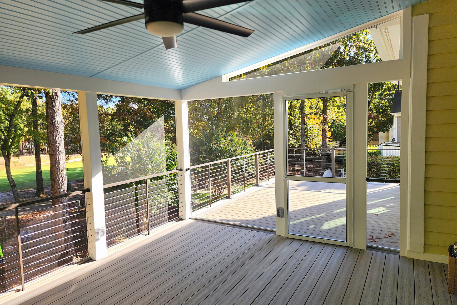 A screened-in porch with light wood decking, a blue ceiling fan, white support beams, and a view of trees.