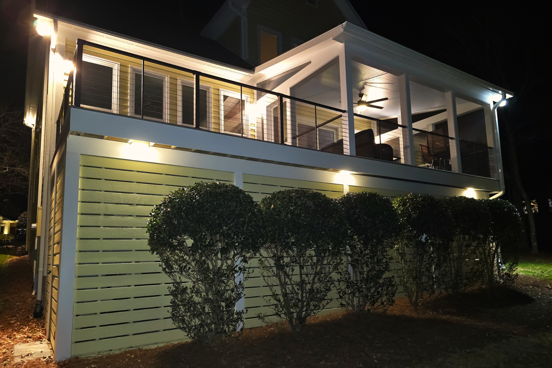 A two-story yellow house at night, featuring a lit upper deck, a screened porch, and bushes illuminated by exterior lights.