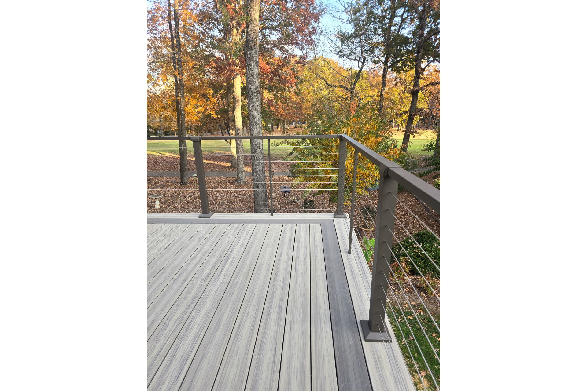 A high-angle view of a grey deck with a metal cable railing overlooking a backyard with autumn trees.