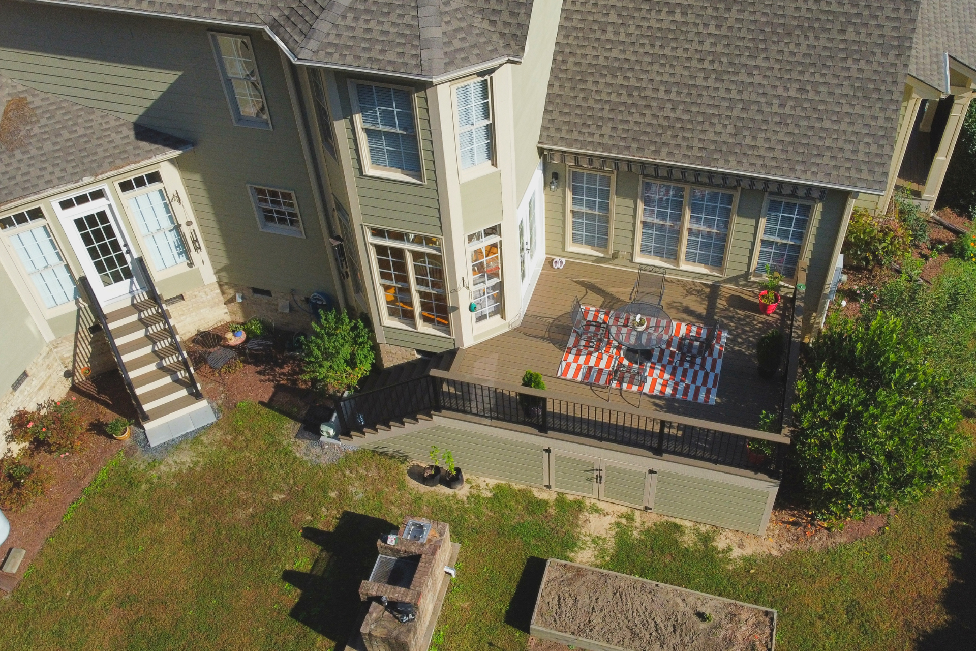 An aerial view of a tan two-story house with a wooden deck, outdoor seating, and a backyard with a small brick structure.