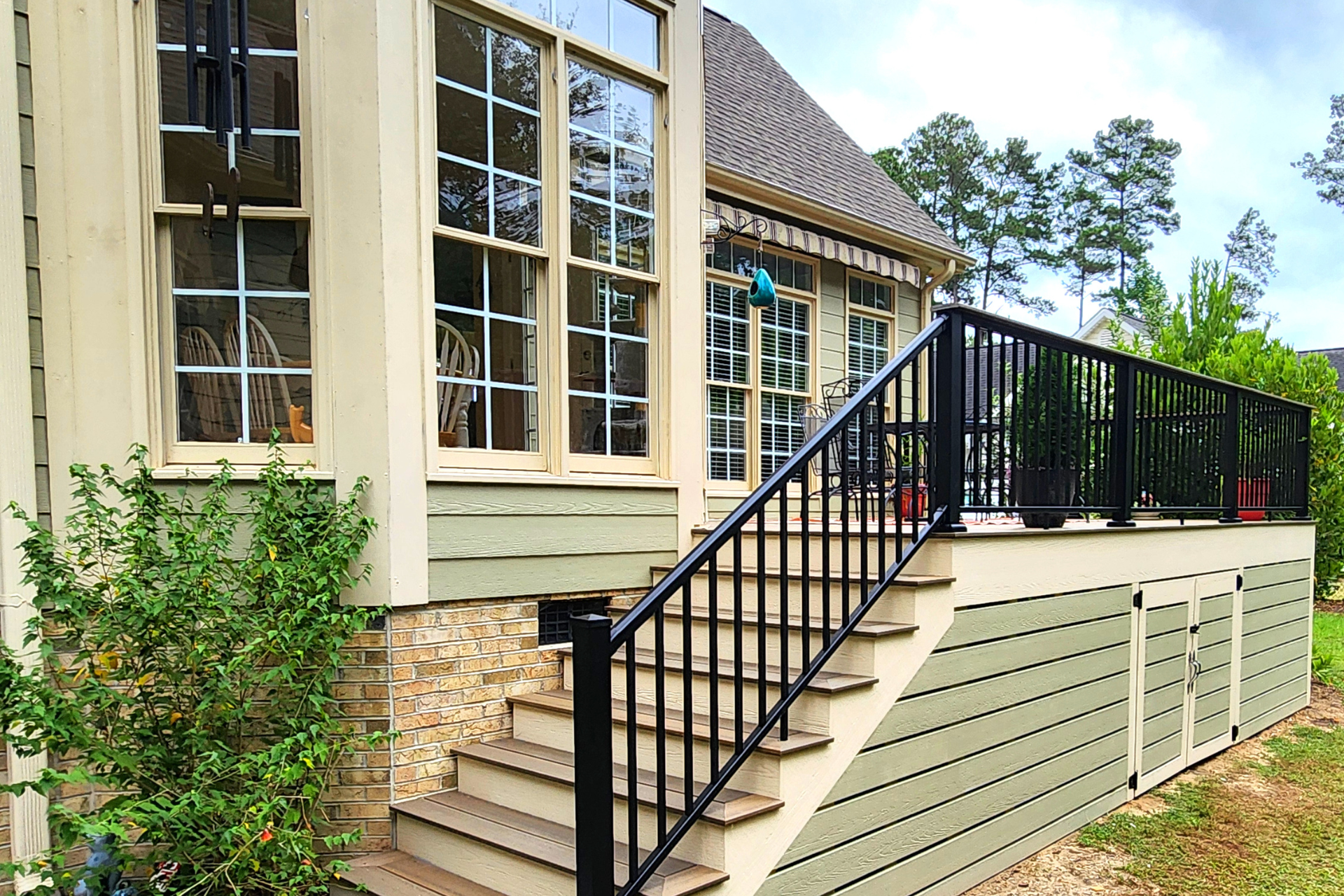 A beige house exterior with a wooden deck, black railings, and a staircase leading to a landscaped yard.