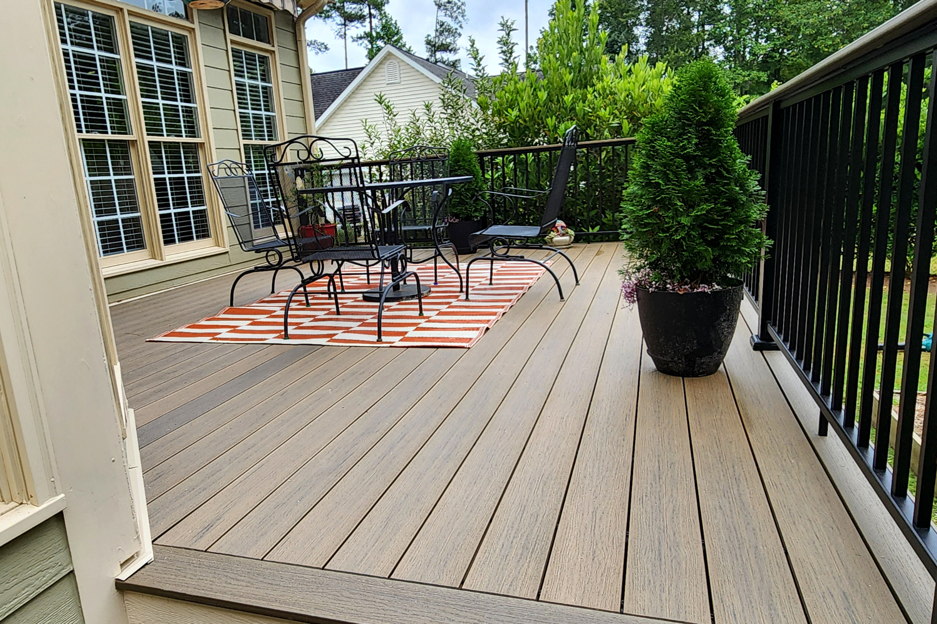 A wooden deck with a table, chairs, an orange-patterned rug, and a potted evergreen plant against a house wall.