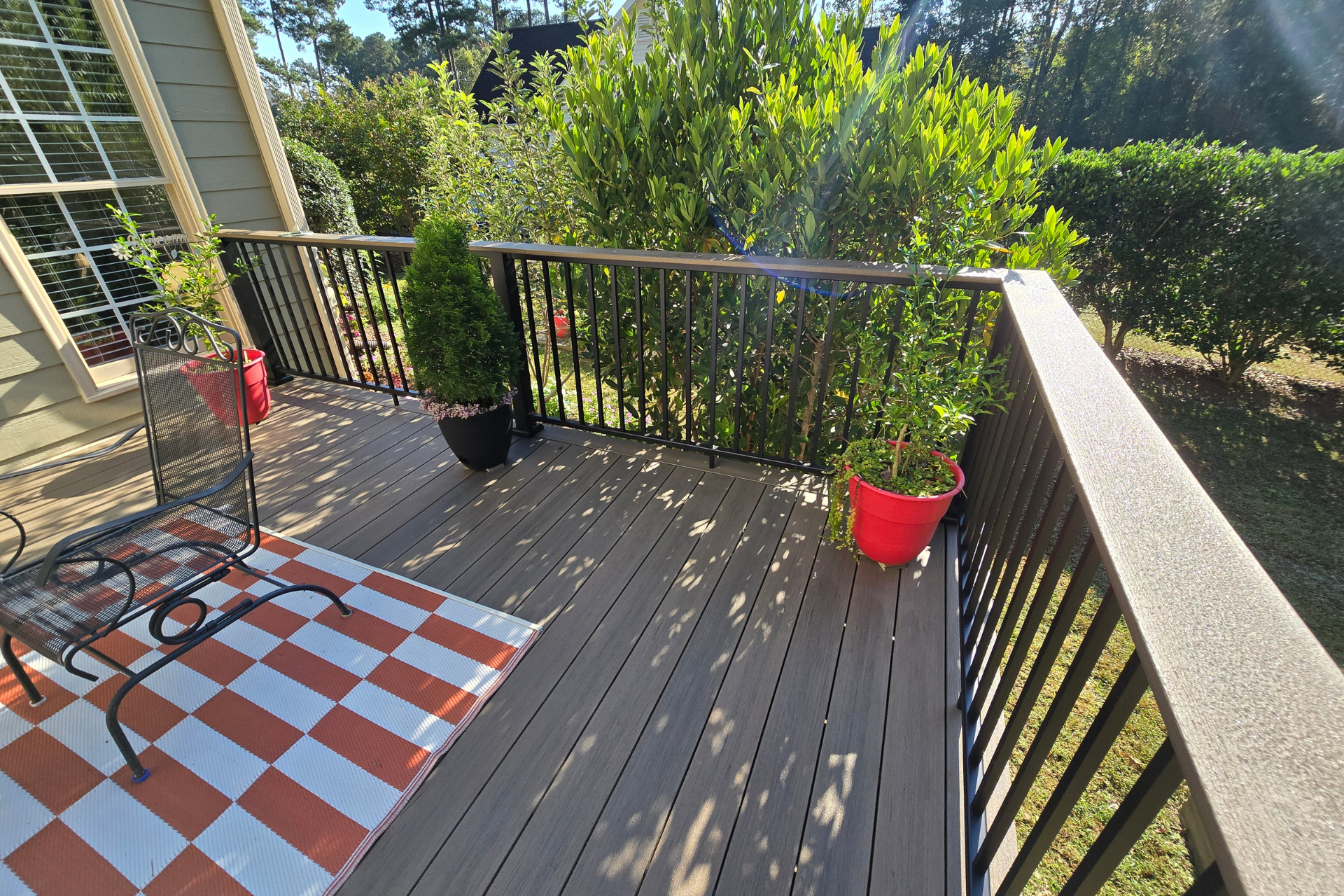 A wooden deck with black metal railings, a checkered orange and white rug, a metal chair, and two potted plants.