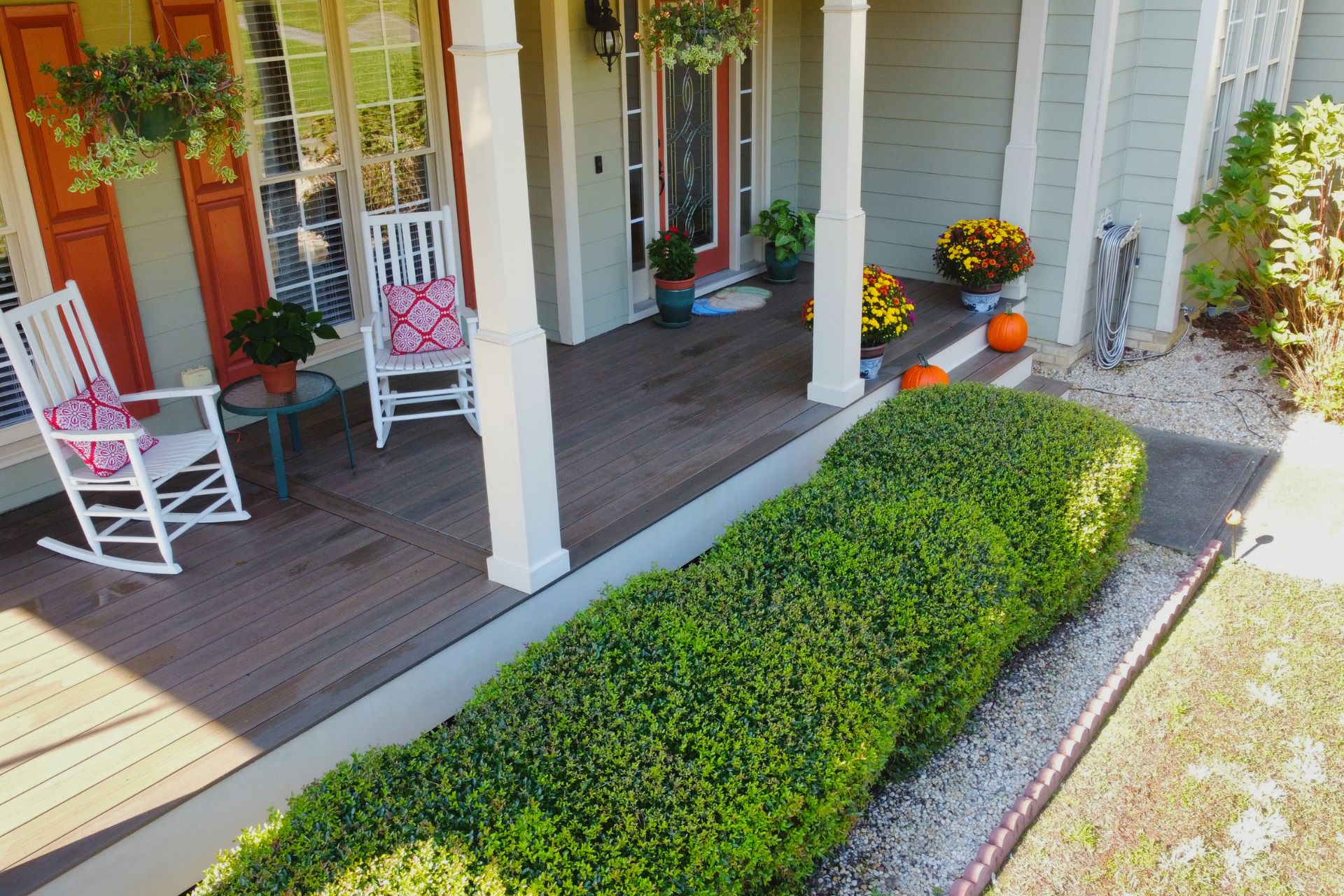 A covered wooden porch features two white rocking chairs, potted plants, and autumn decorations including pumpkins.