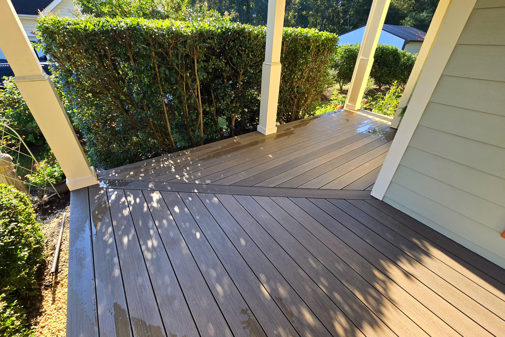 A light-brown wood-plank deck with a diagonal design, bordered by tall green hedges and white porch columns.