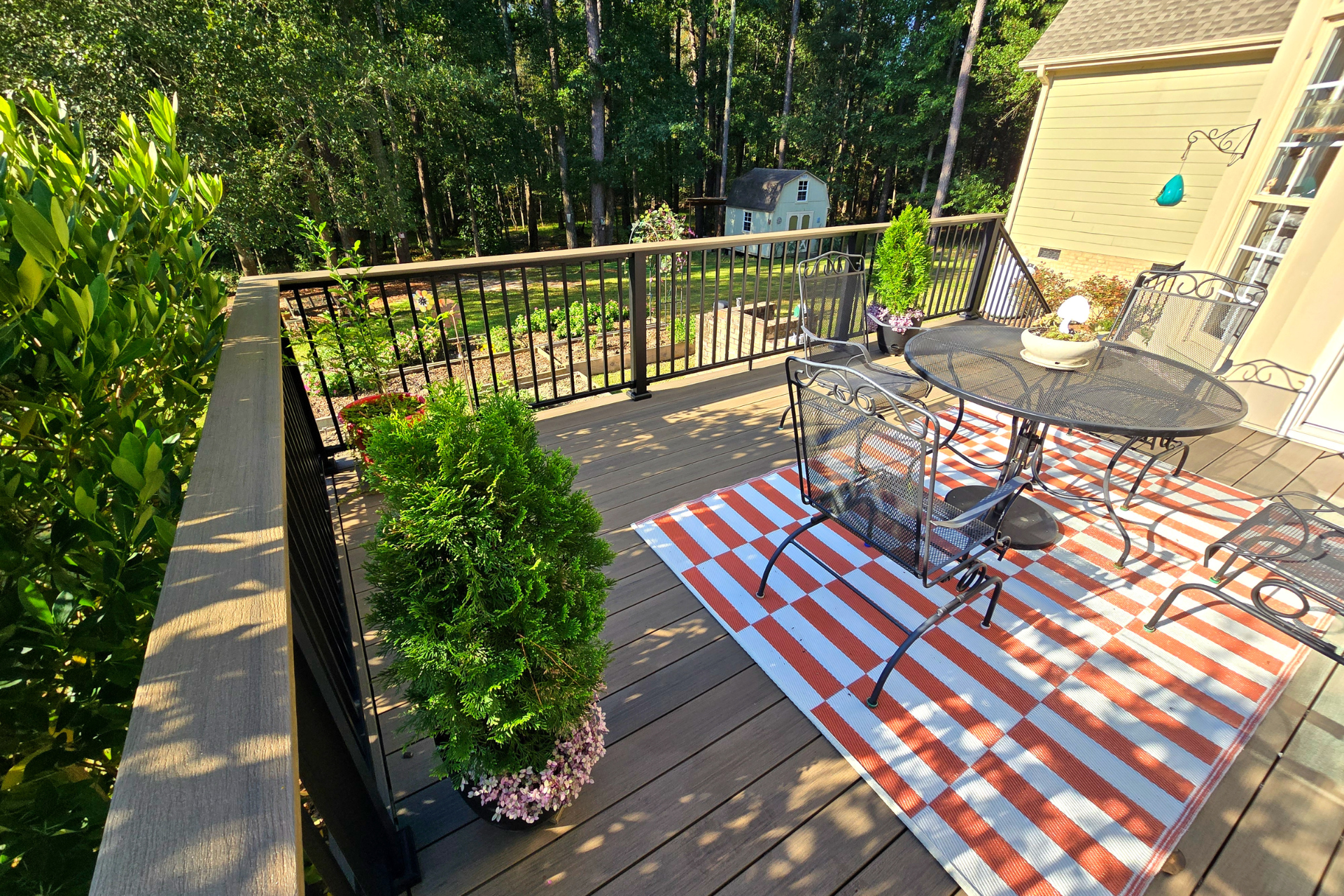 A wooden deck with a table, chairs, and a red-and-white striped rug, overlooking a lush green yard and trees.