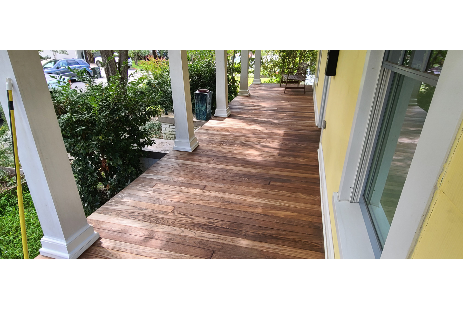 A long, wood-planked porch with white pillars along a yellow house exterior, leading toward a yard with green foliage.