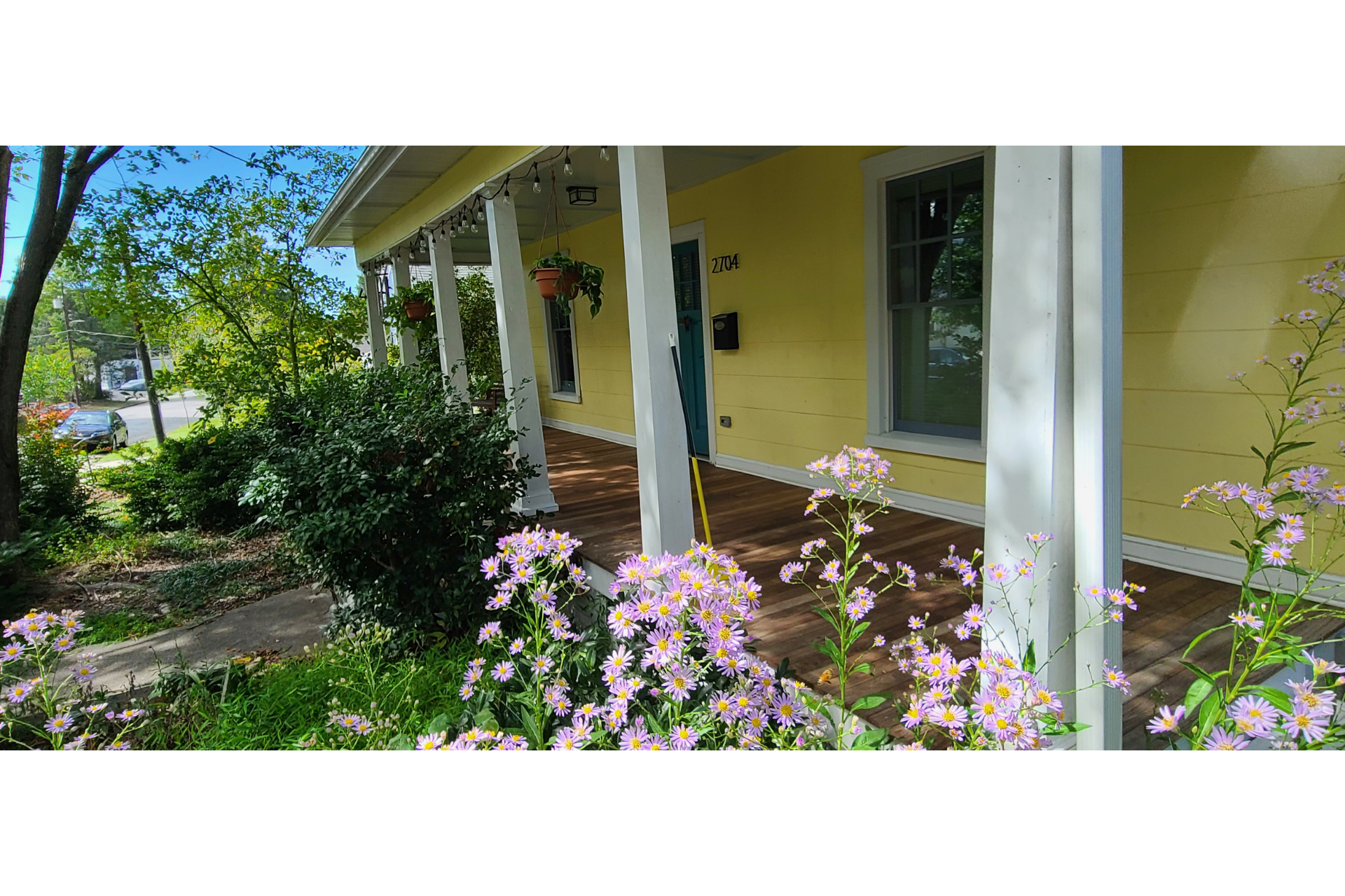 A yellow house with a covered porch, white pillars, and purple flowers in the foreground.