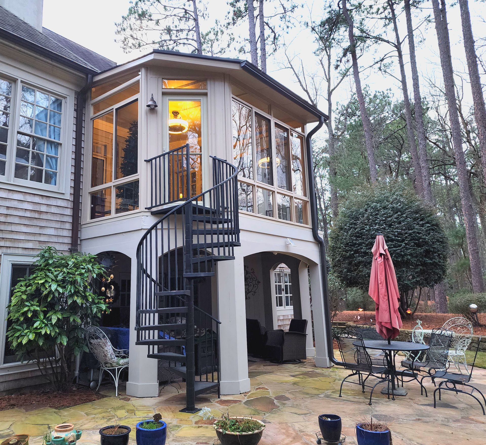 A two-story home exterior featuring a second-story screened porch connected to a paved patio by a black spiral staircase.