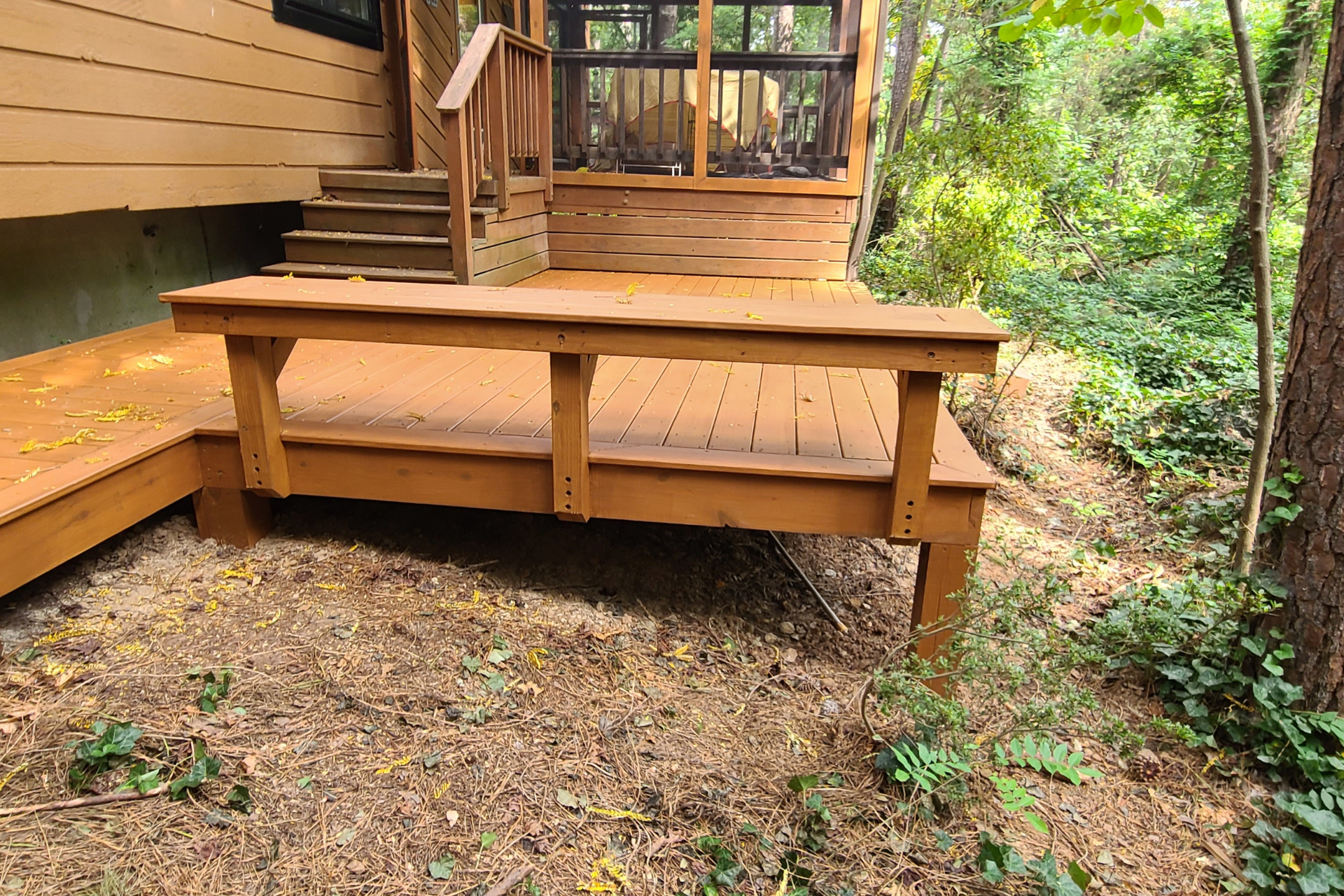 A wooden deck attached to a house features a built-in bench, steps leading to a screened porch, and a surrounding yard.