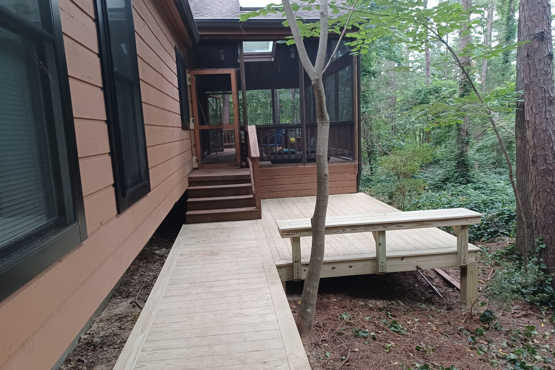 A wooden deck with a built-in bench leading to stairs and a screened-in porch on the side of a tan house in a wooded area.
