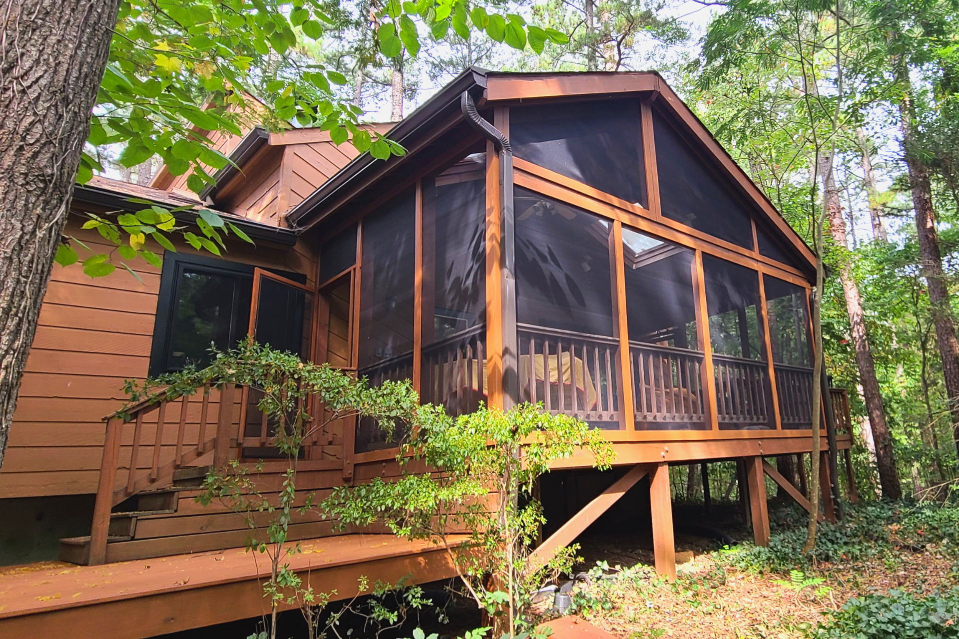 A brown wooden house with a screened-in porch and deck surrounded by trees in a wooded area.