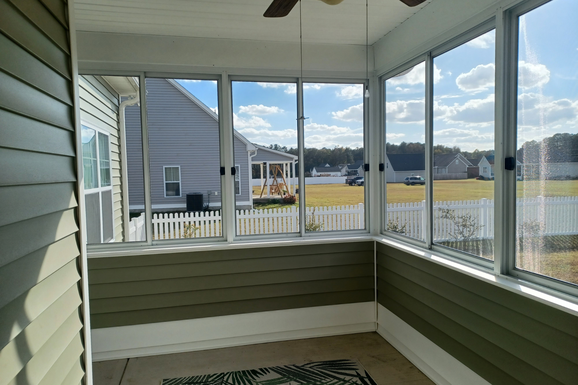 A bright sunroom with green horizontal siding, large windows, and a white picket fence visible outside.