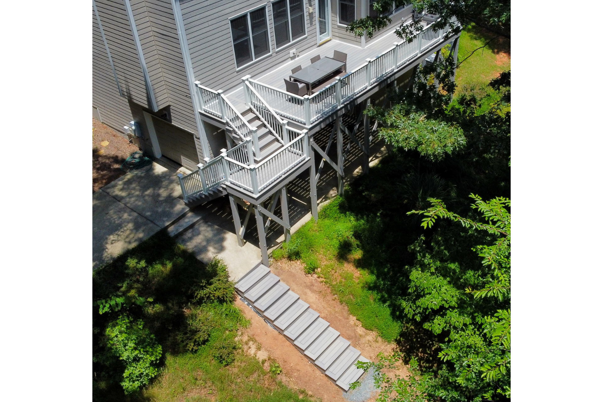 A light gray elevated deck with a staircase leading down to a concrete path in a wooded backyard.