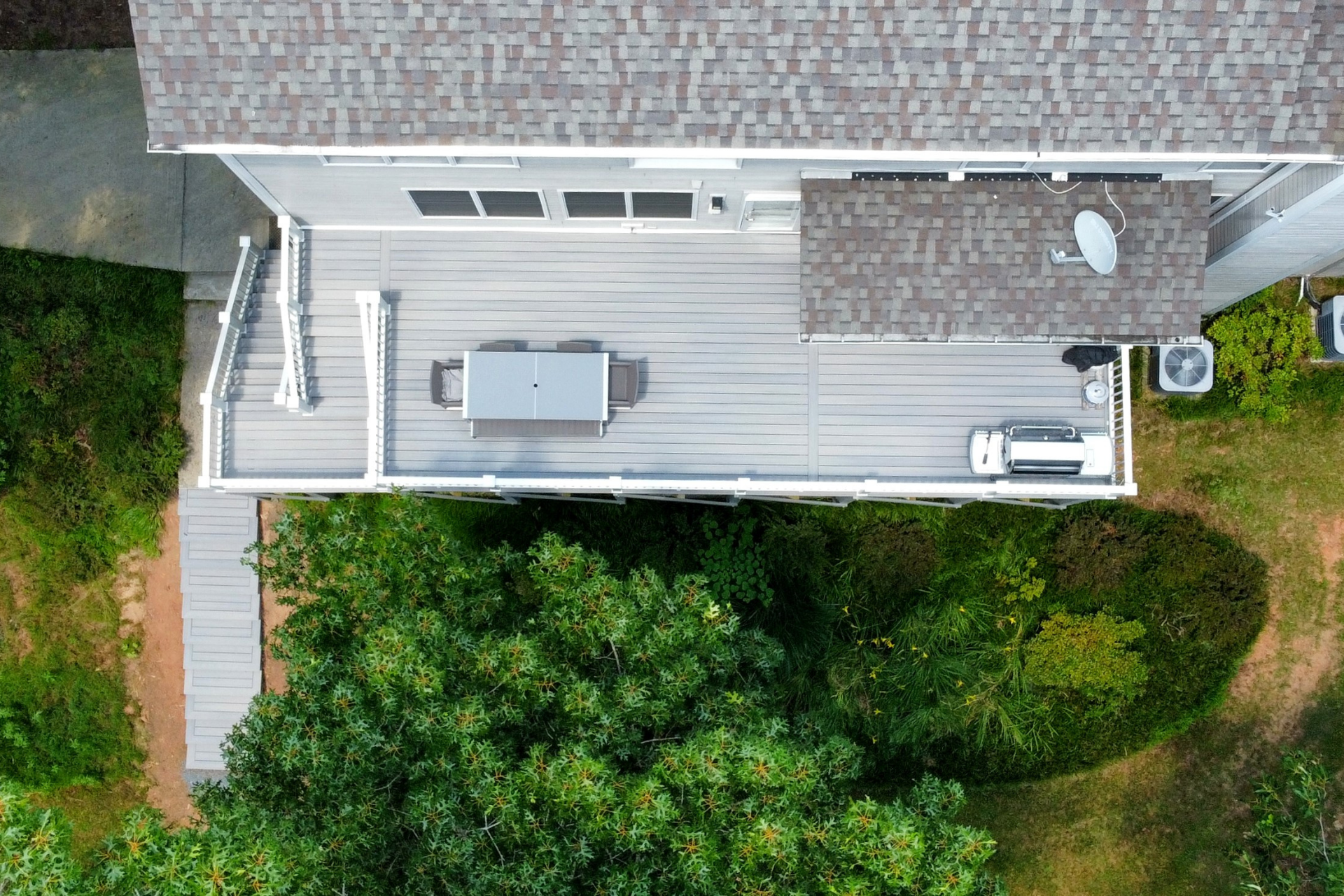 Aerial view of a gray deck attached to a house, featuring a table, grill, and surrounding trees.