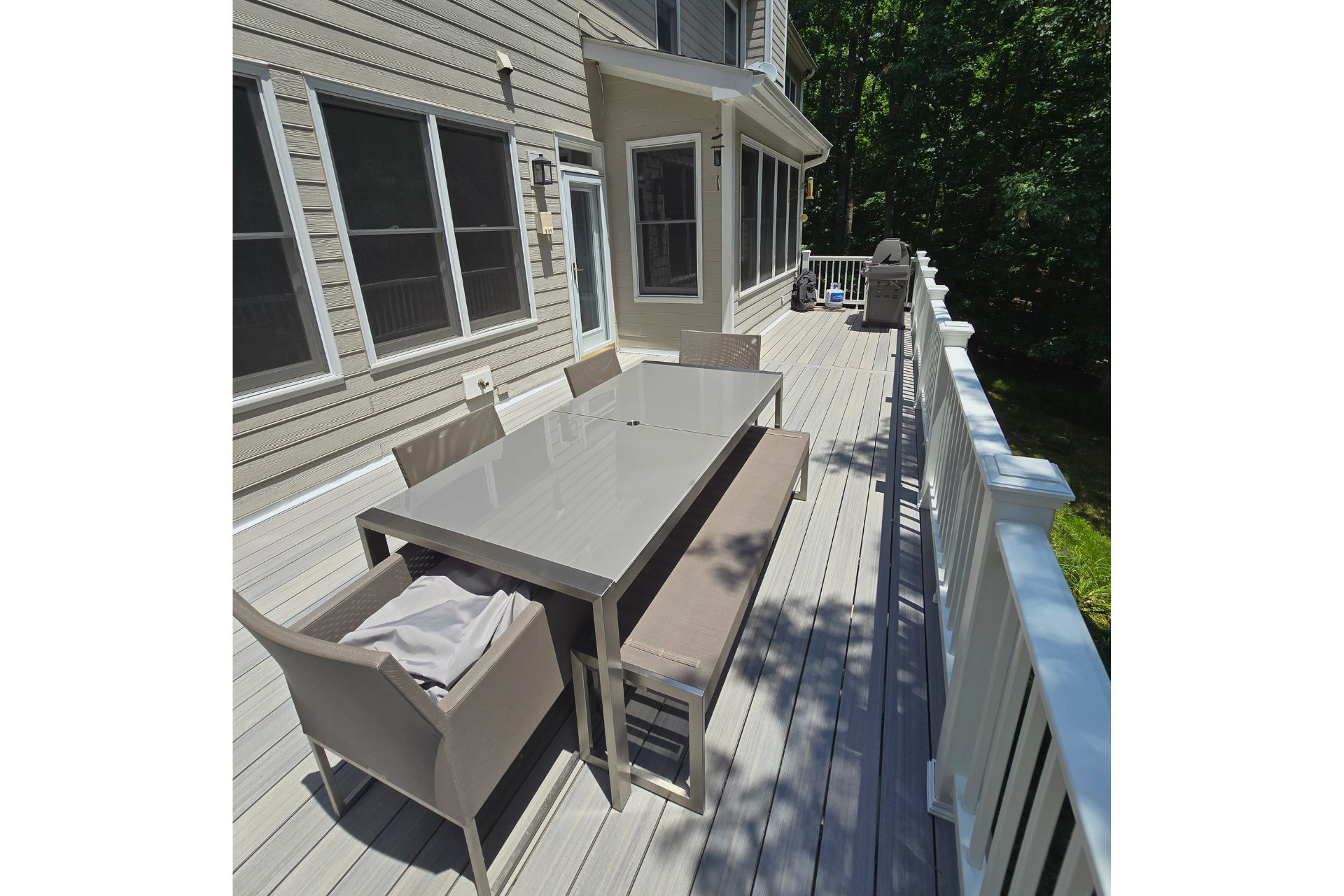 A modern, neutral-colored patio table set with benches and chairs sits on a light-colored wooden deck beside a house.