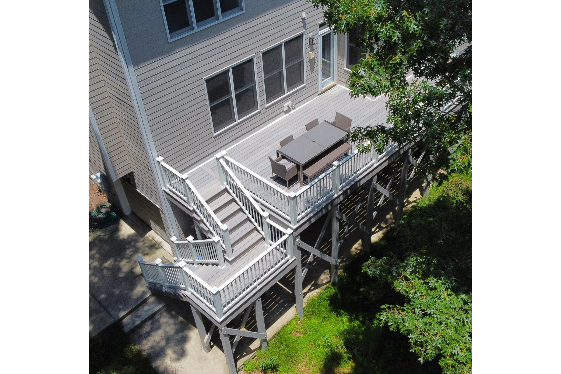 A gray elevated wooden deck with stairs leading to a backyard, featuring a dining set under the shade of nearby trees.