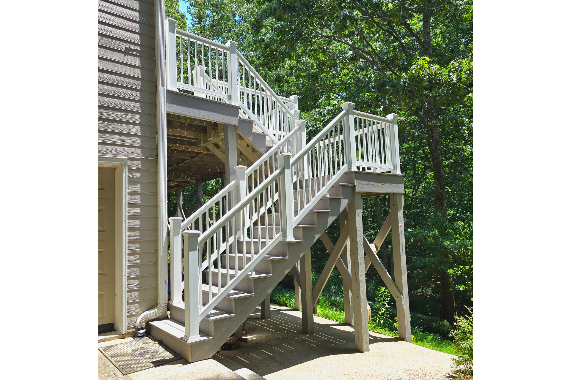White wooden stairs with matching railings lead up to a raised outdoor deck next to a brick house and forest.
