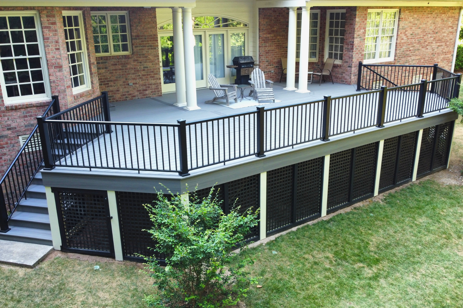 A raised grey composite deck with black metal railings, lattice skirting, and white columns attached to a brick house.