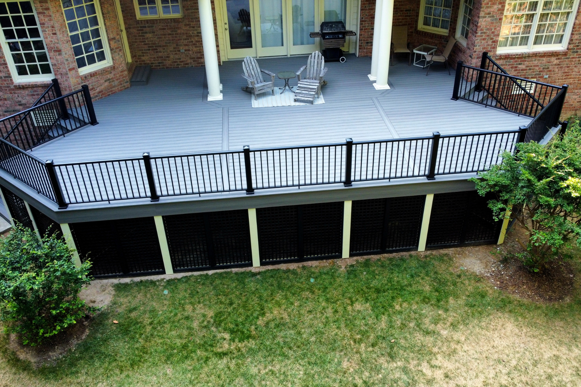 A raised gray deck with black railings and lattice skirting stands outside a brick house with white pillars and chairs.