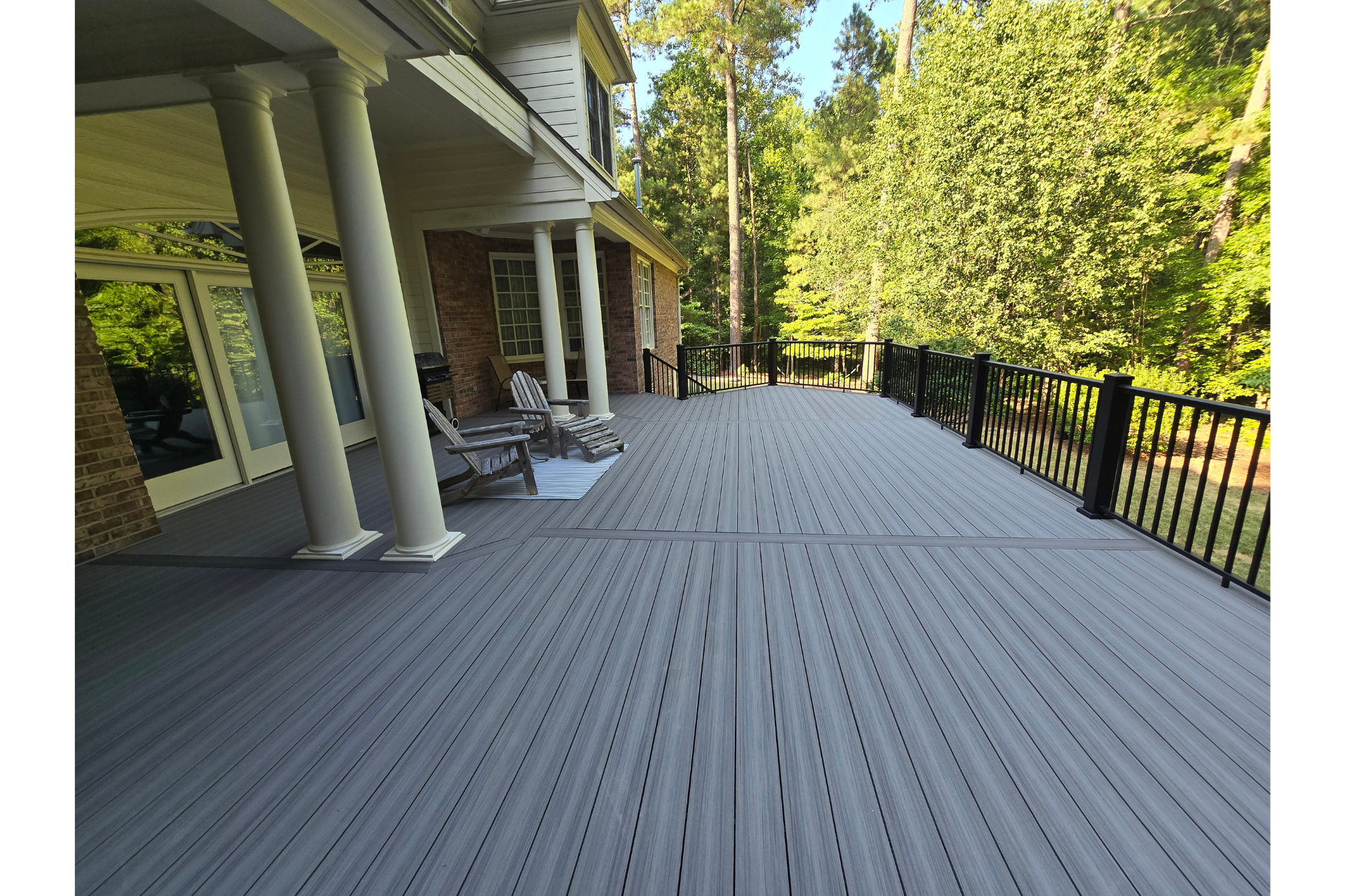 A spacious gray composite deck with a black railing, overlooking a wooded backyard by a house with white pillars.