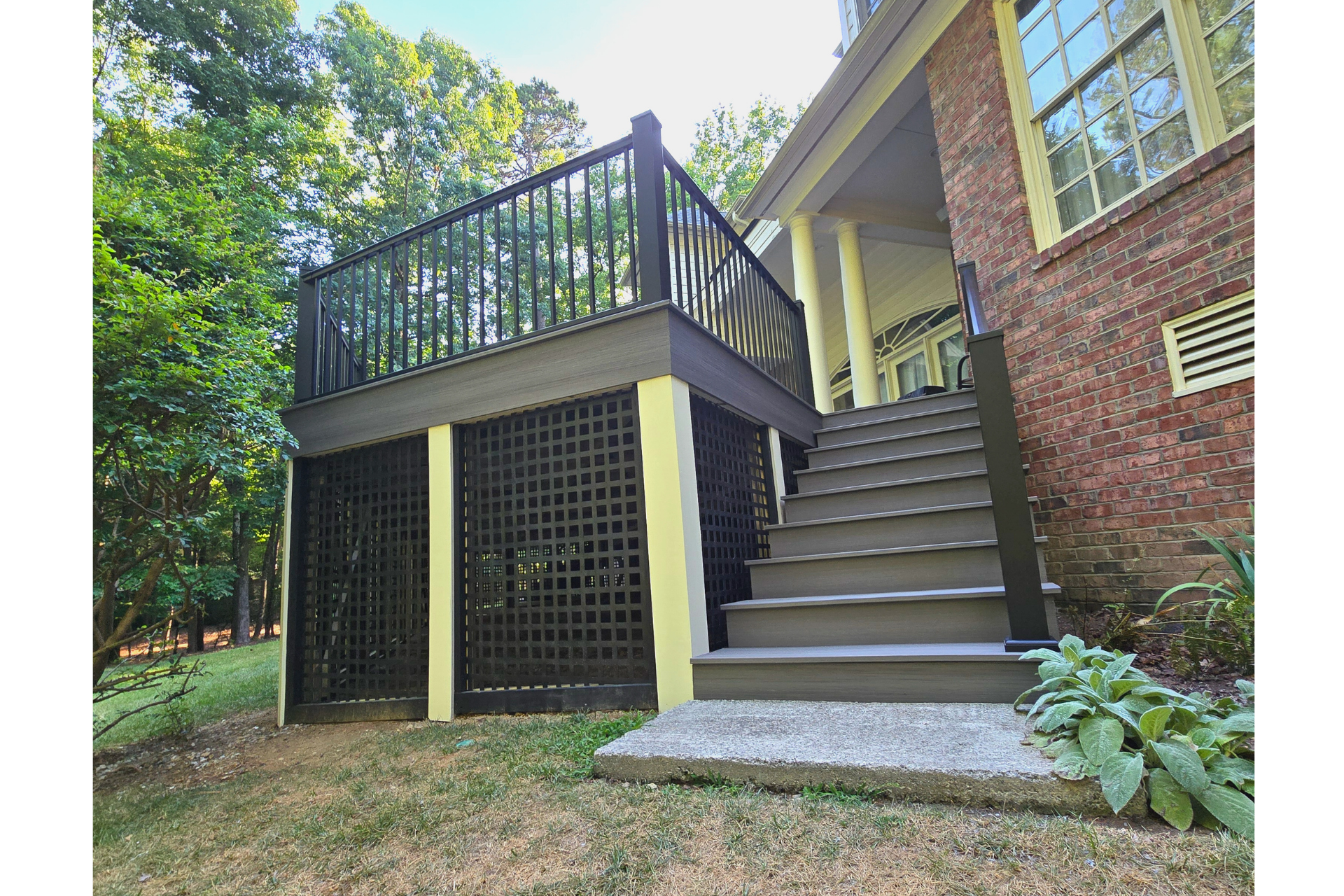 A raised deck with dark railings and lattice skirting, accessed by wide brown stairs against a brick house exterior.