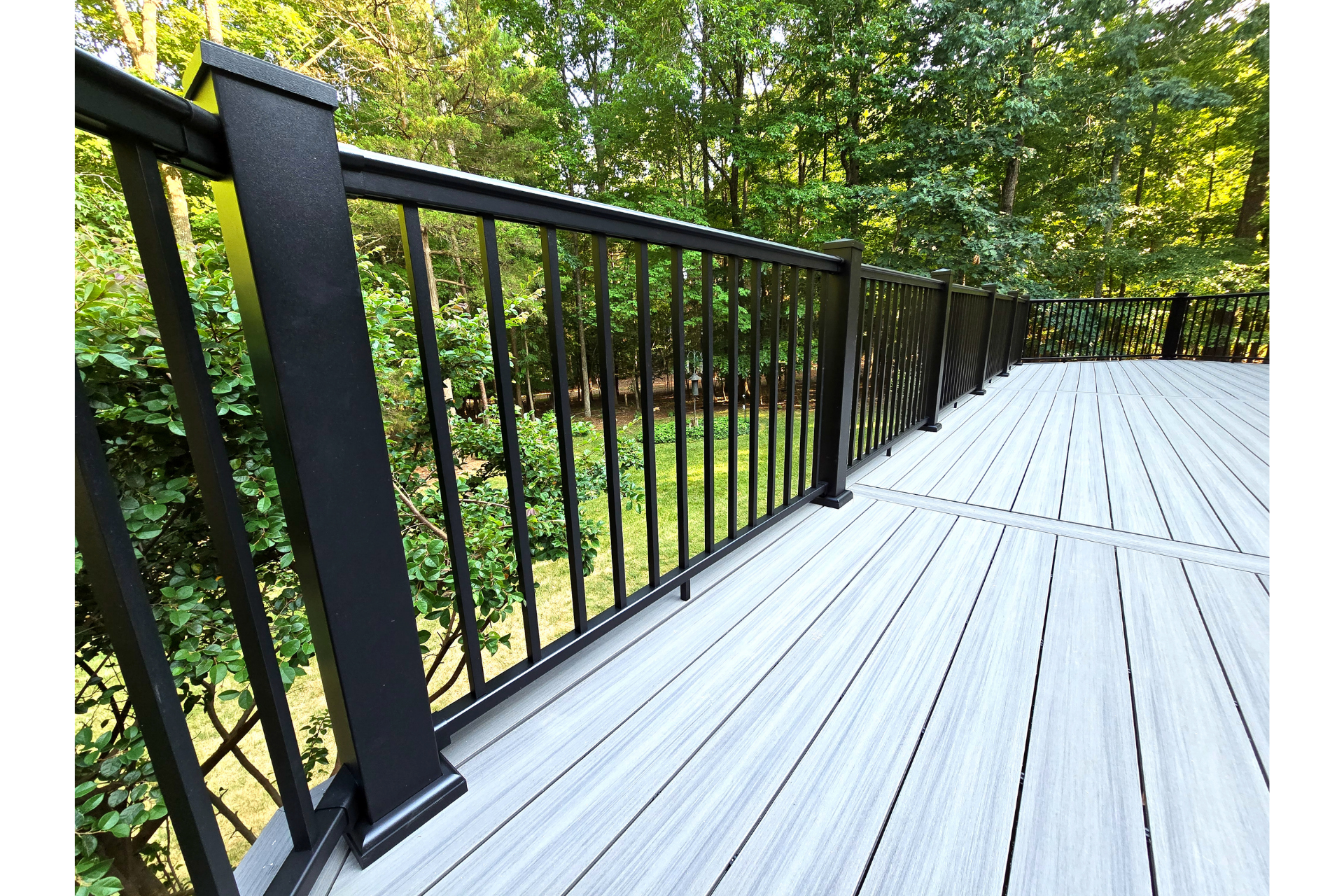A matte black metal railing borders a light gray, wood-grain composite deck surrounded by lush green trees.