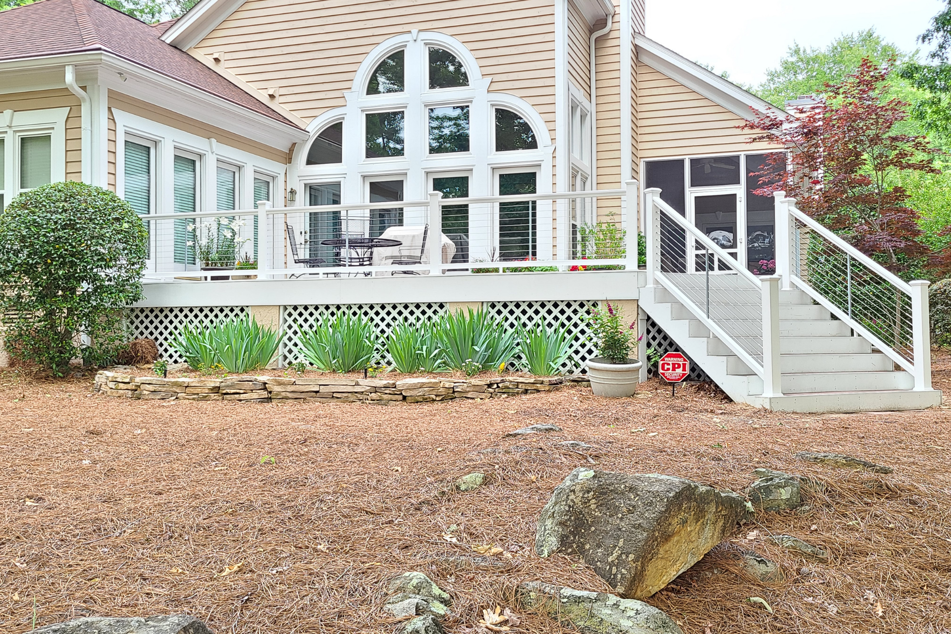 A beige house with a large, elevated white deck, staircase, and lattice skirting, surrounded by a yard with wood mulch.