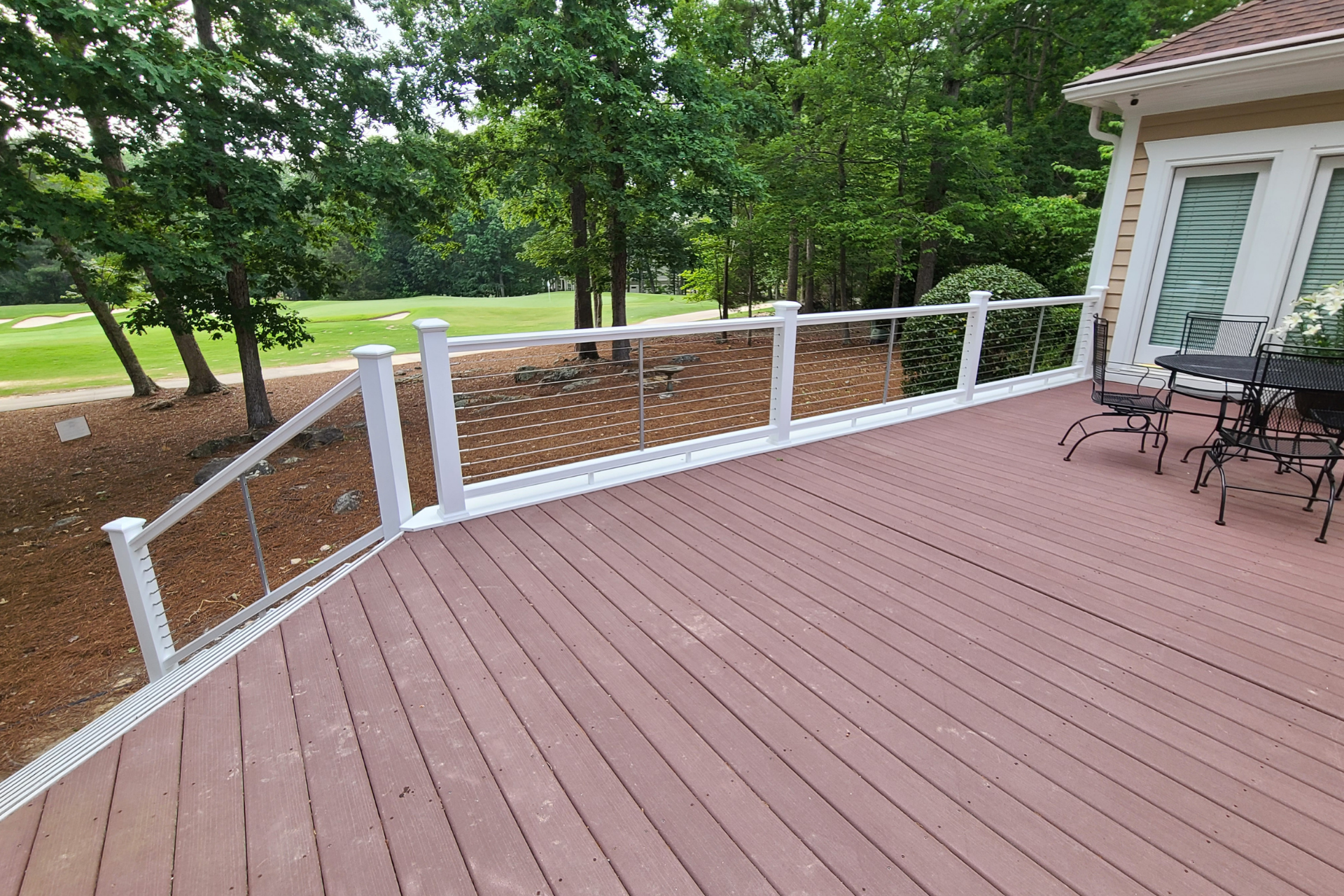 A reddish-brown composite deck with white railings and metal cable infill, overlooking a grassy area and trees.