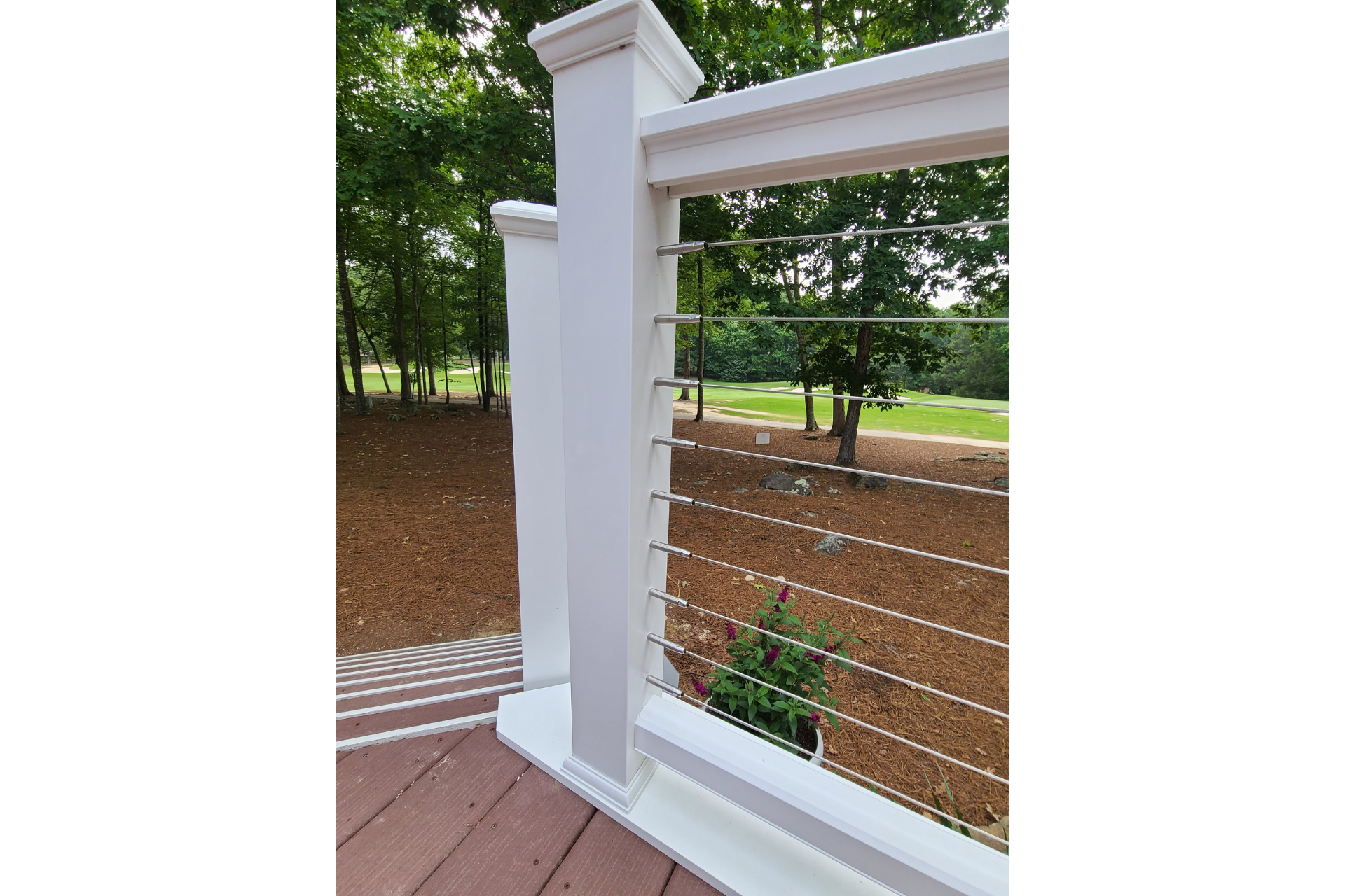 A white railing system with thin horizontal metal cables, installed on a deck overlooking a wooded backyard.