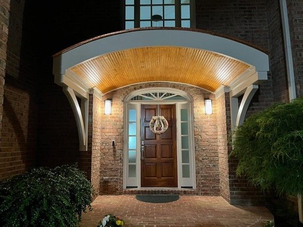 Brick home entryway with a wooden door, illuminated porch lights, and a hanging wreath.