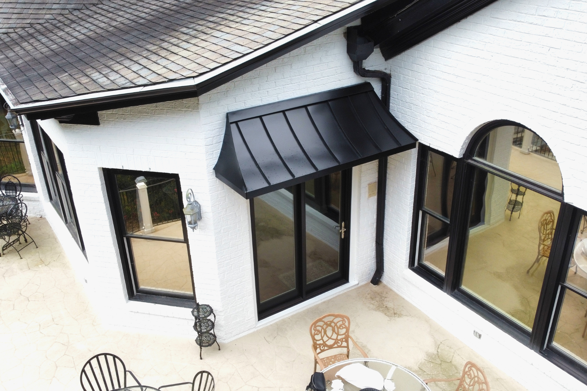 A high-angle view of a white brick patio with black-trimmed windows, a black door awning, and outdoor furniture.