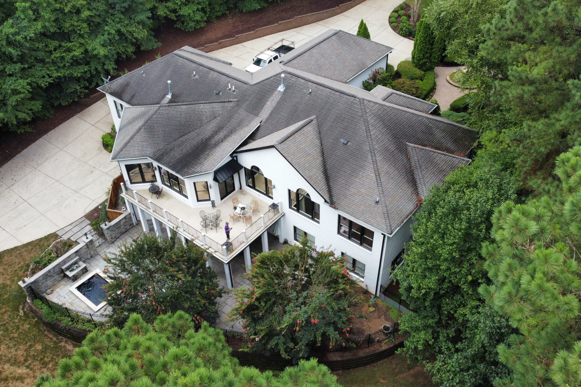 Aerial view of a white, multi-story suburban home with a dark roof, back deck, and small pool, surrounded by trees.