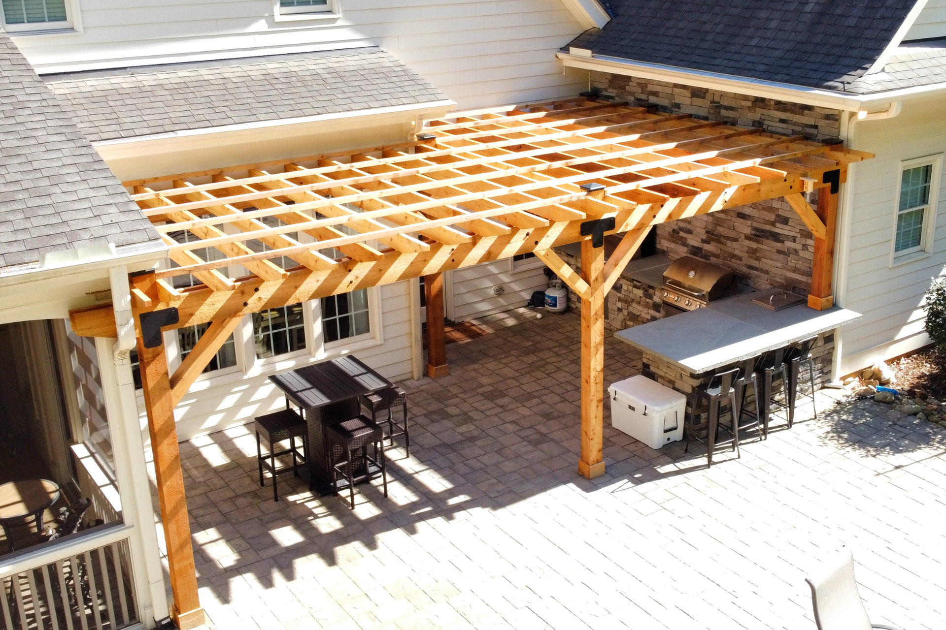 A high-angle view of a backyard patio featuring a wooden pergola, an outdoor dining set, and a built-in stone grill station.