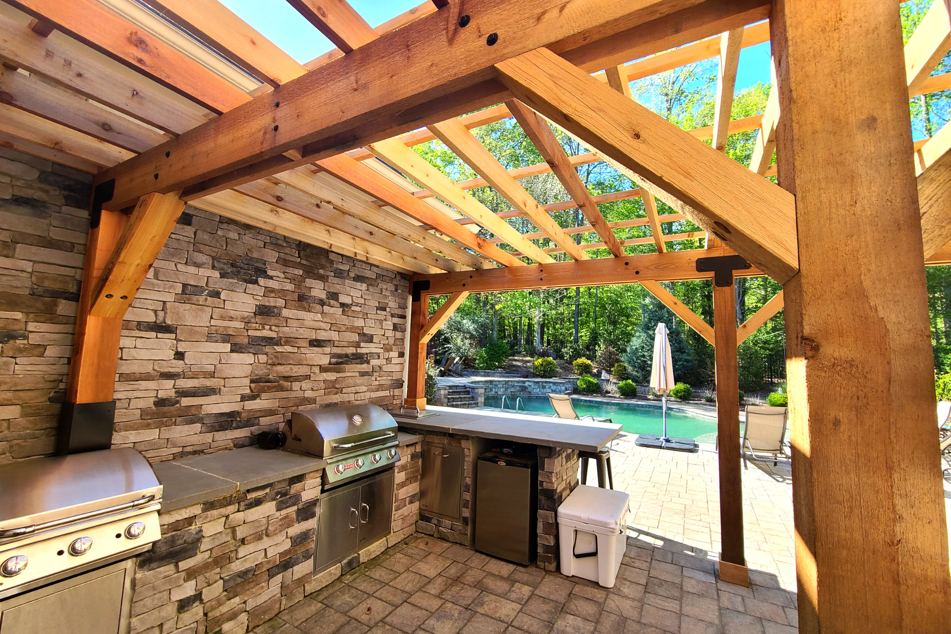 Outdoor kitchen with stainless steel appliances, stacked stone walls, and a timber pergola overlooking a swimming pool.